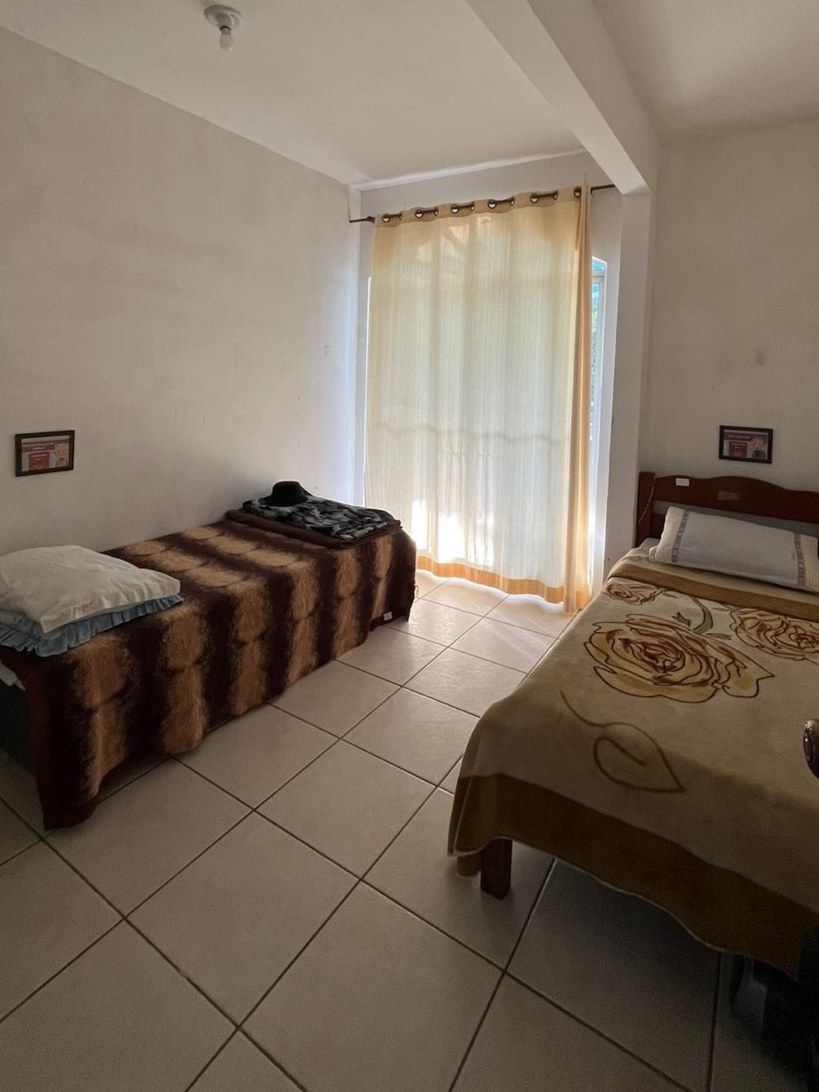 Quarto duplo com duas camas, ambiente modesto, limpo e iluminado por uma porta-janela